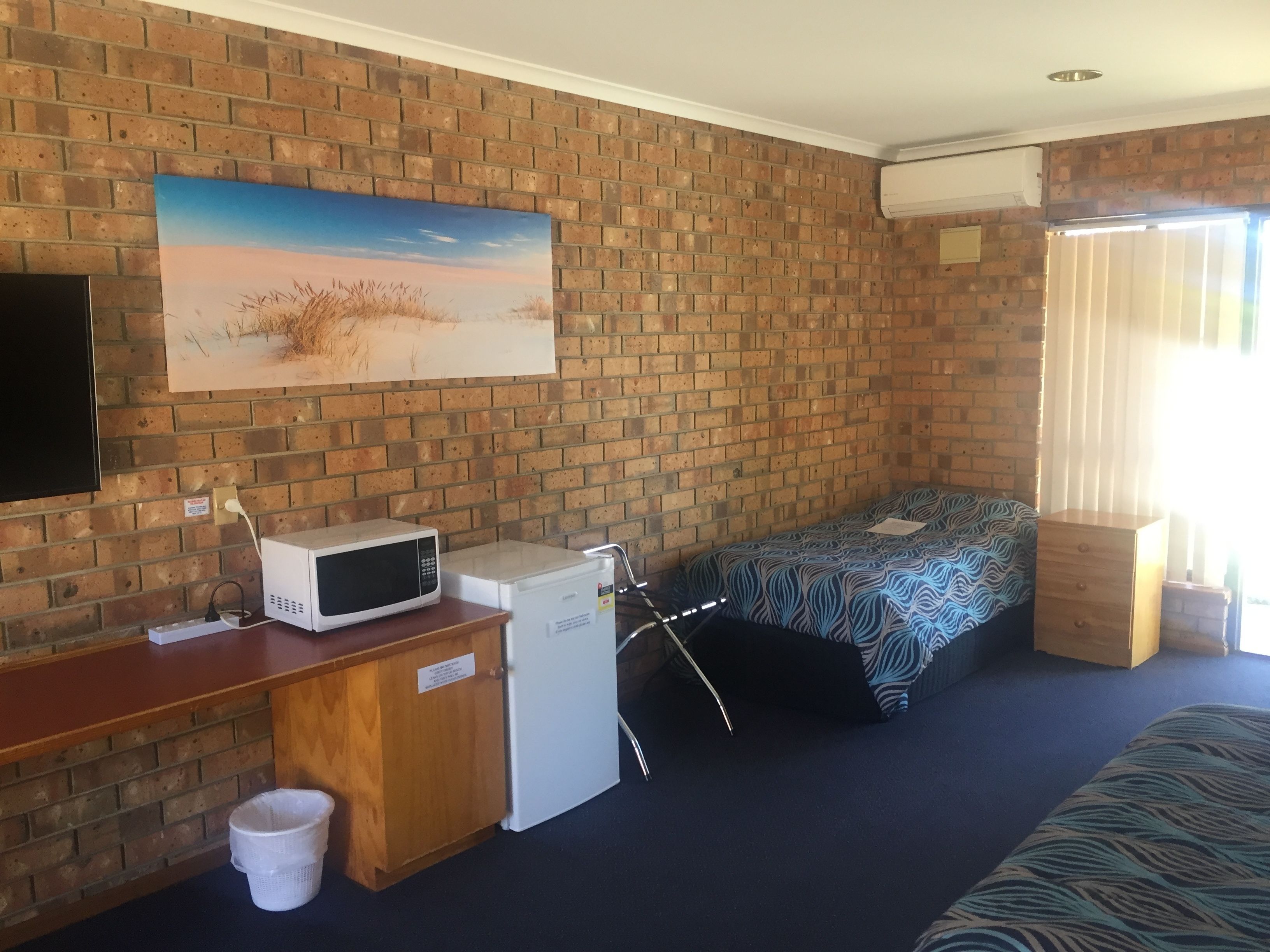 A room with a brick wall, a painting of a landscape, a bed with a zebra print cover, a microwave, a mini fridge, a wooden desk, and a trash bin.
