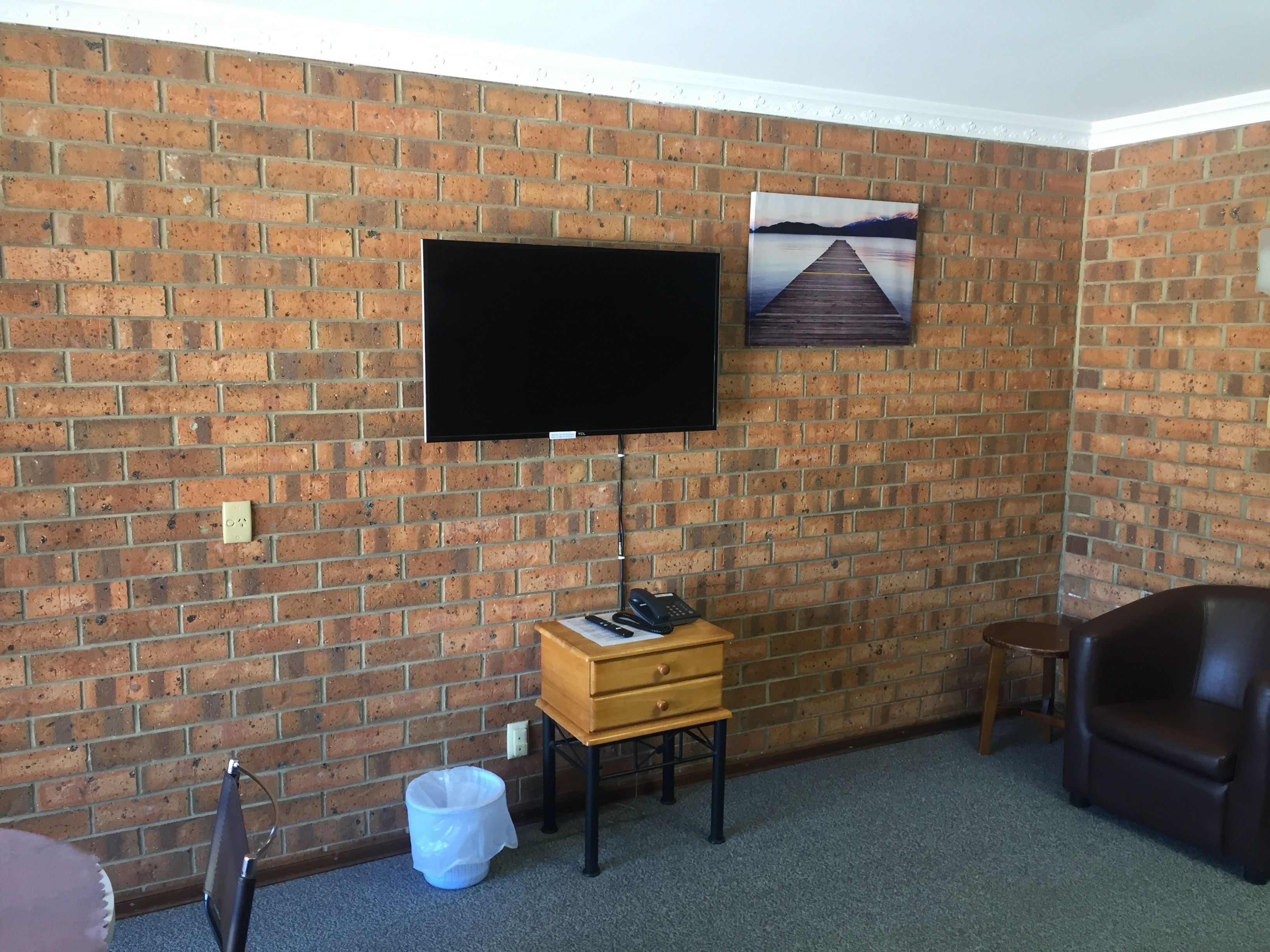 A room with a brick wall, a flat-screen TV mounted above a wooden desk, a telephone, and a painting of a pier.