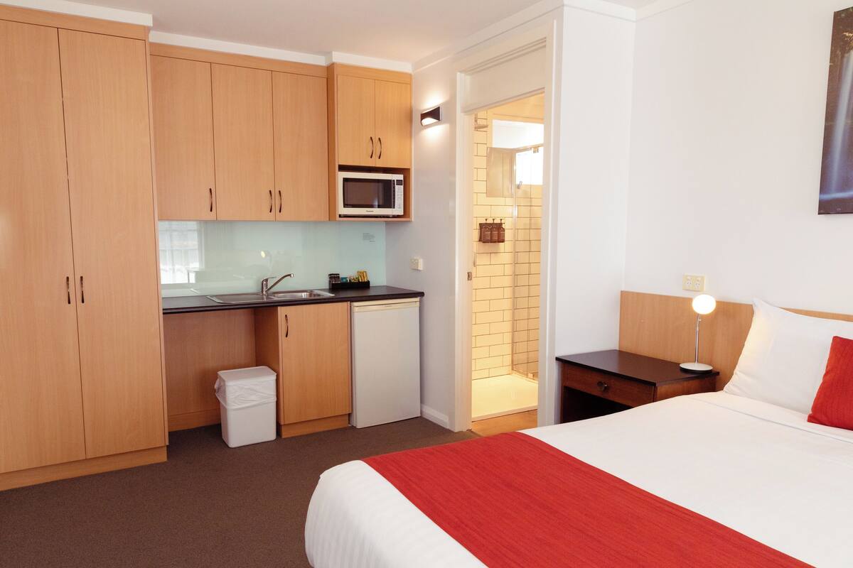 A hotel room with a bed, a nightstand, a desk, a microwave, a sink, and a trash bin.