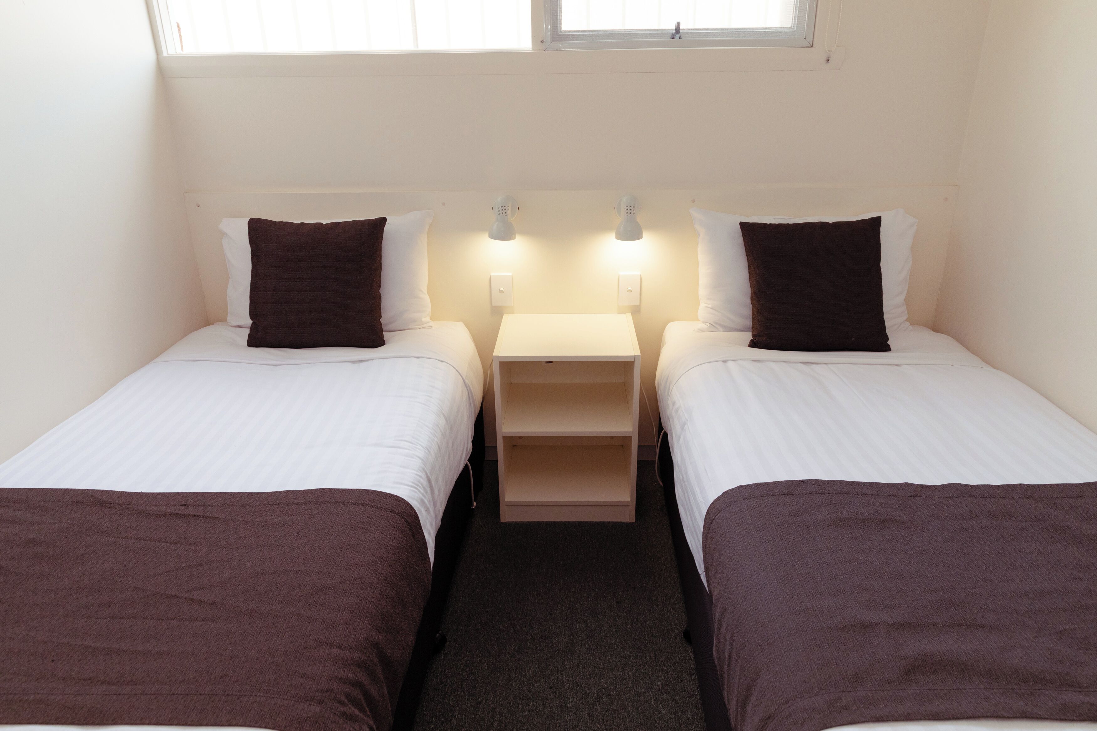 Habitación doble con dos camas individuales, cada una con una almohada y una manta. Pequeño mesita de noche con lámpara entre las camas.