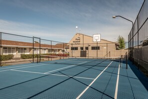 An outdoor tennis court with a net, surrounded by chain-link fencing, and a building labeled "The Pool Heated 20°C + Perfect" in the background.