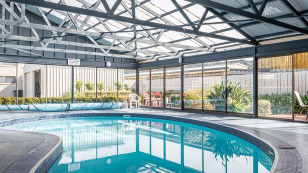 Una piscina interior con techo de cristal, rodeada por un patio con sillas y plantas.