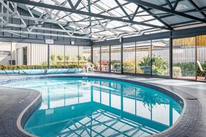 An indoor swimming pool with a glass ceiling, surrounded by a patio area with chairs and plants.