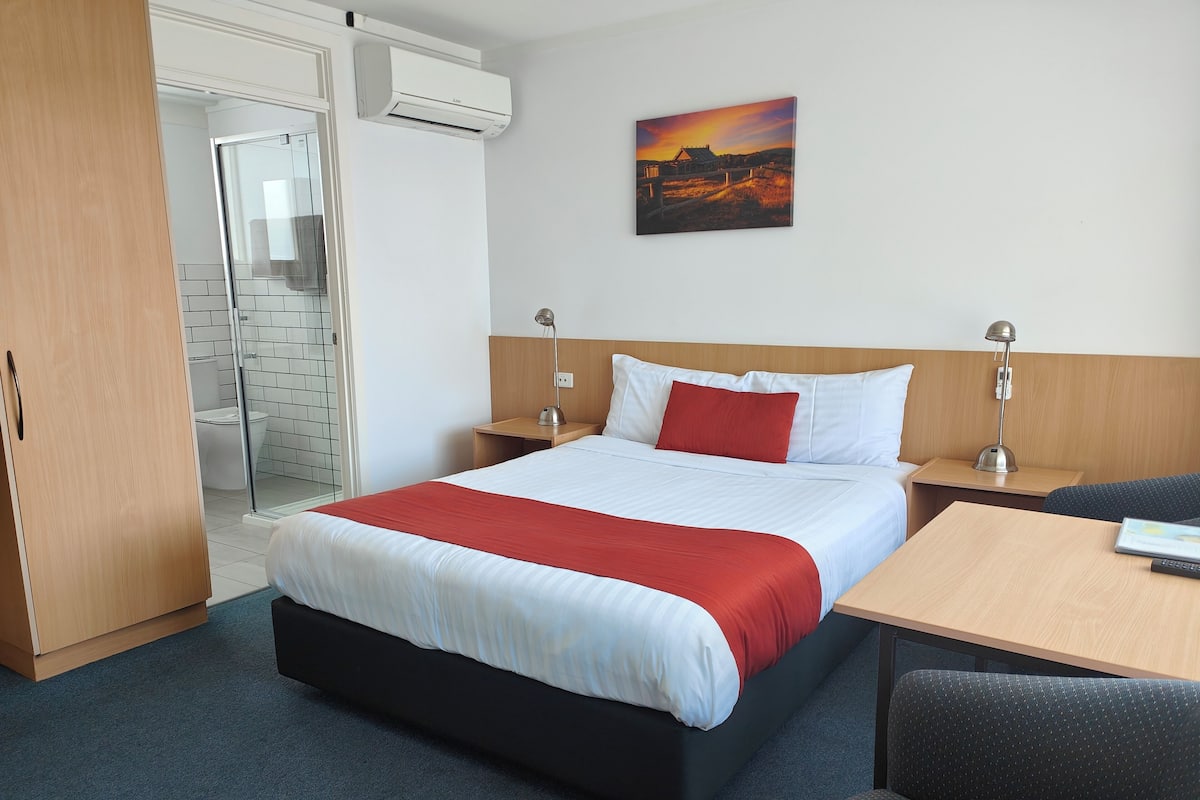 A hotel room with a bed, a desk, a chair, a painting, and an air conditioning unit.