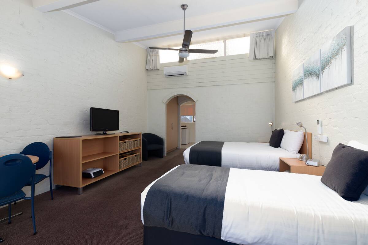 A hotel room with two beds, a desk, a chair, a television, and a ceiling fan.