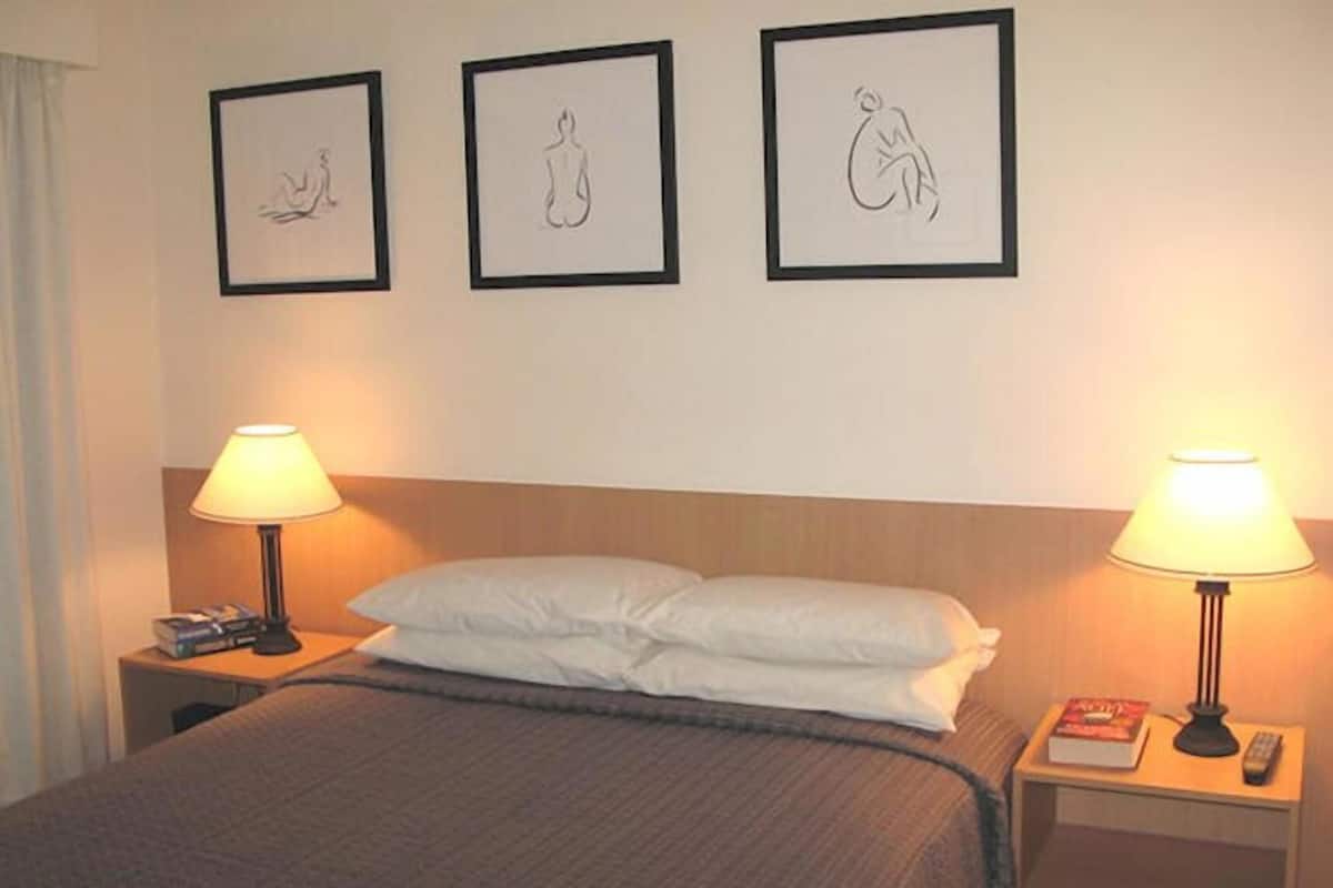 A hotel room with a bed, two bedside lamps, a nightstand with a book and remote, and three framed wall art pieces.