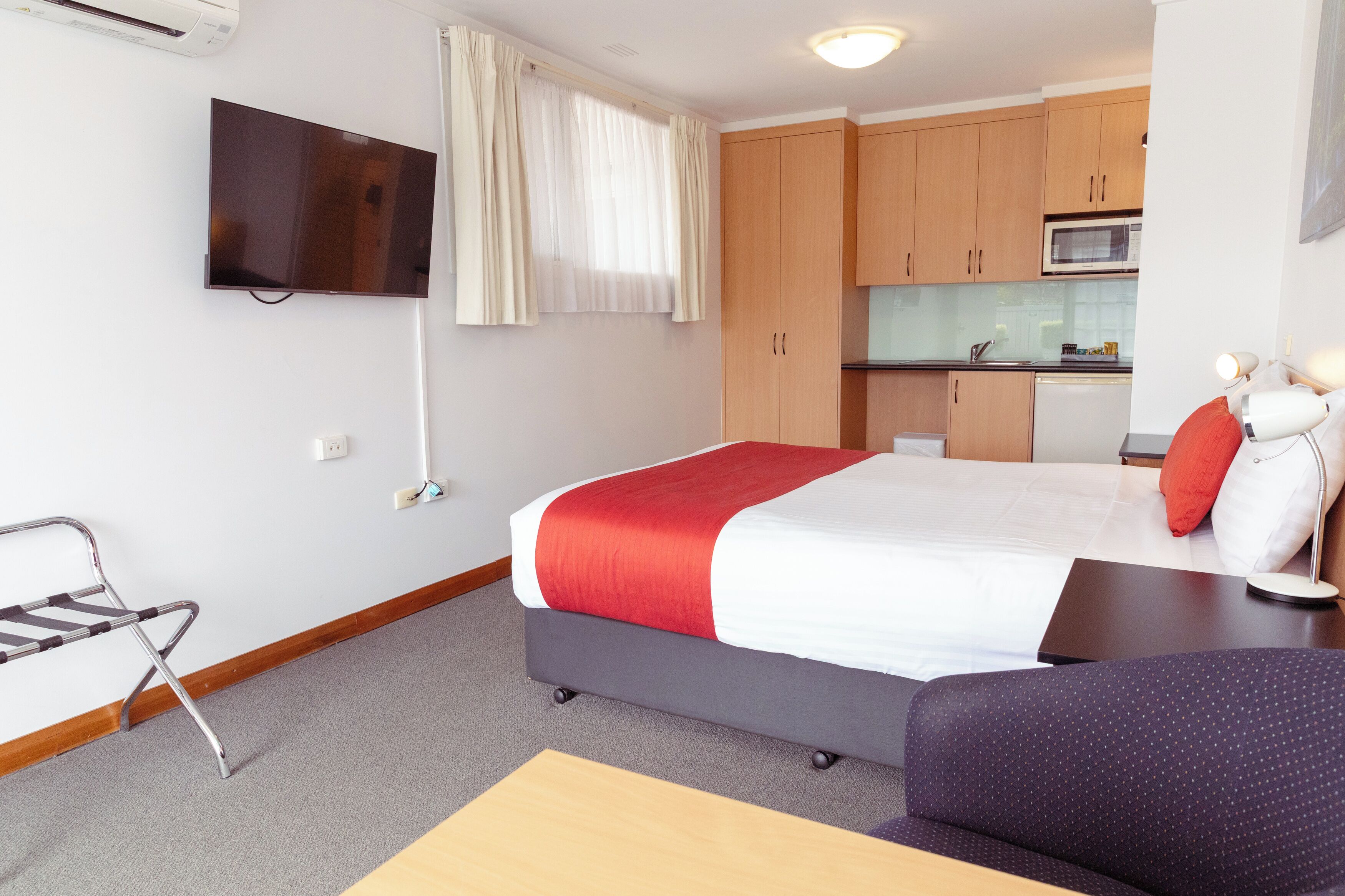 A hotel room with a bed, a TV mounted on the wall, a kitchenette, and a seating area with a sofa and a chair.