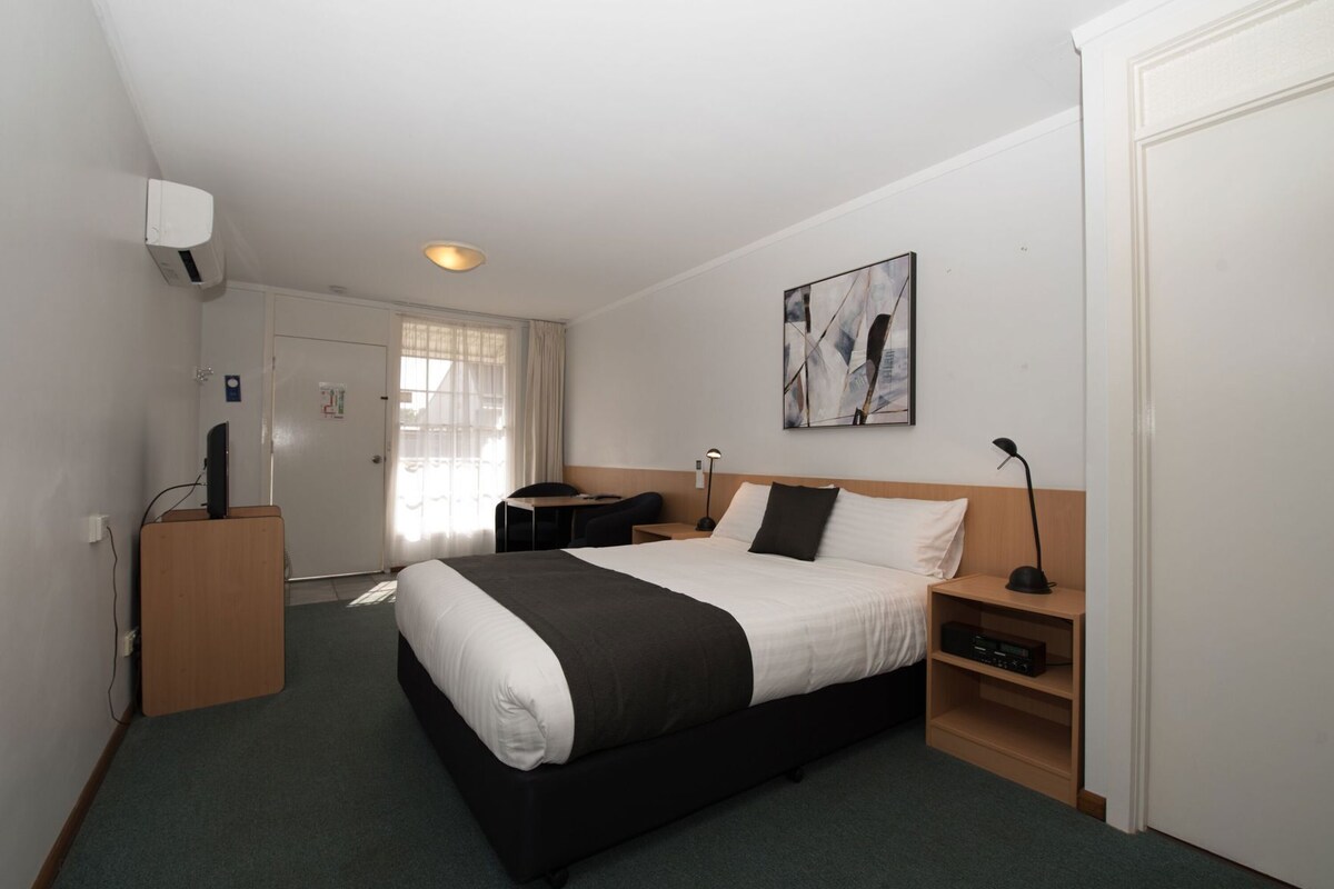 Una habitación de hotel con cama, mesita de noche, escritorio, silla y un cuadro en la pared.