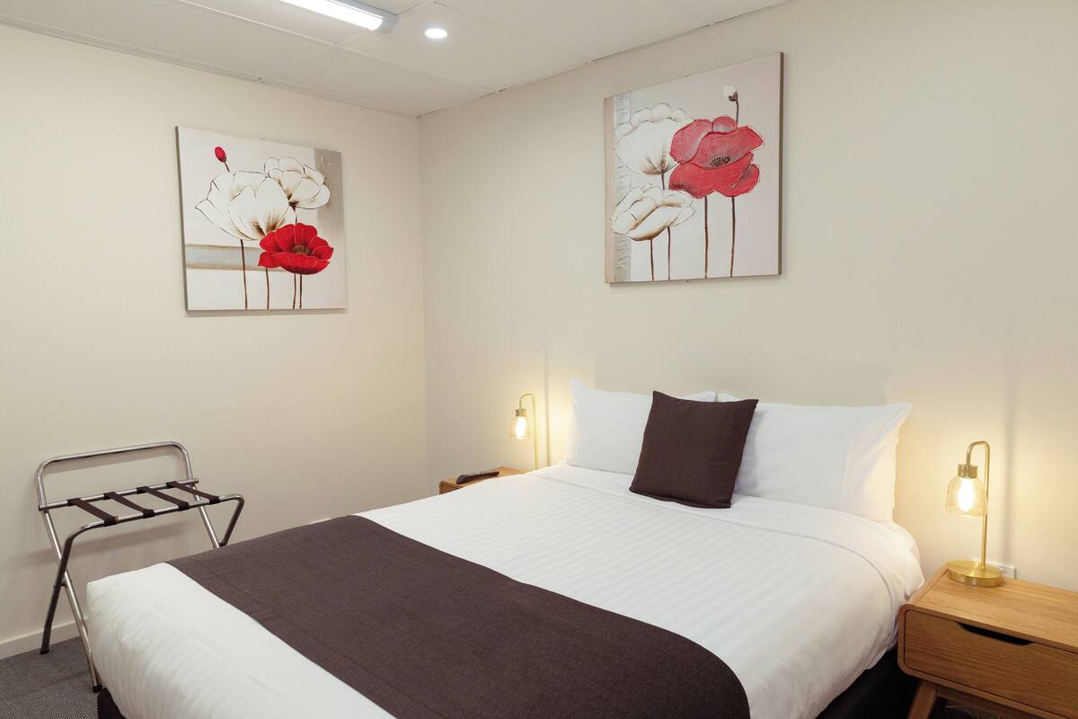 A neatly made bed with a brown blanket, two bedside lamps, and two wall-mounted paintings of flowers.