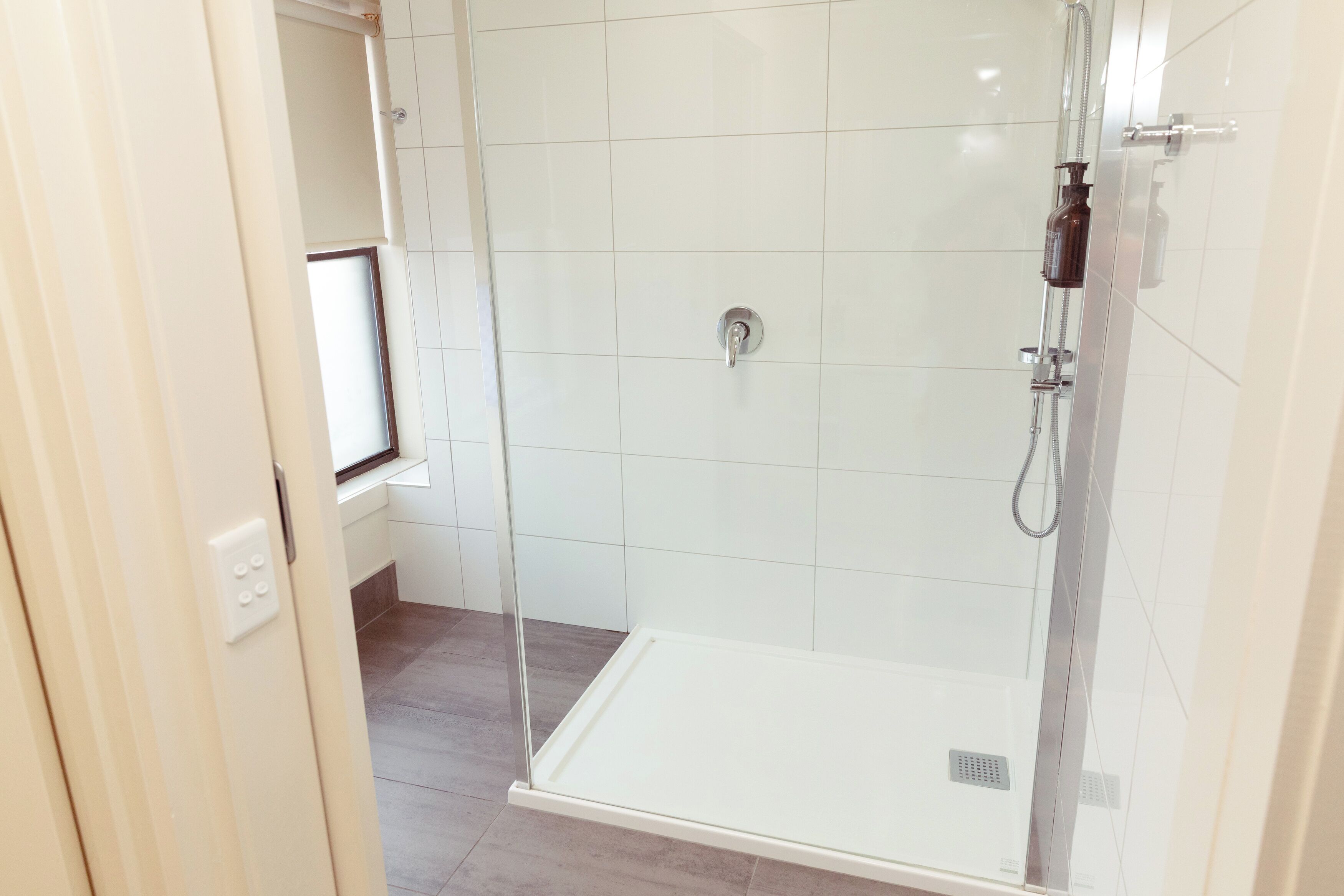 Zona de ducha con mampara de vidrio, pared con azulejos blancos, panel de control y ventana.