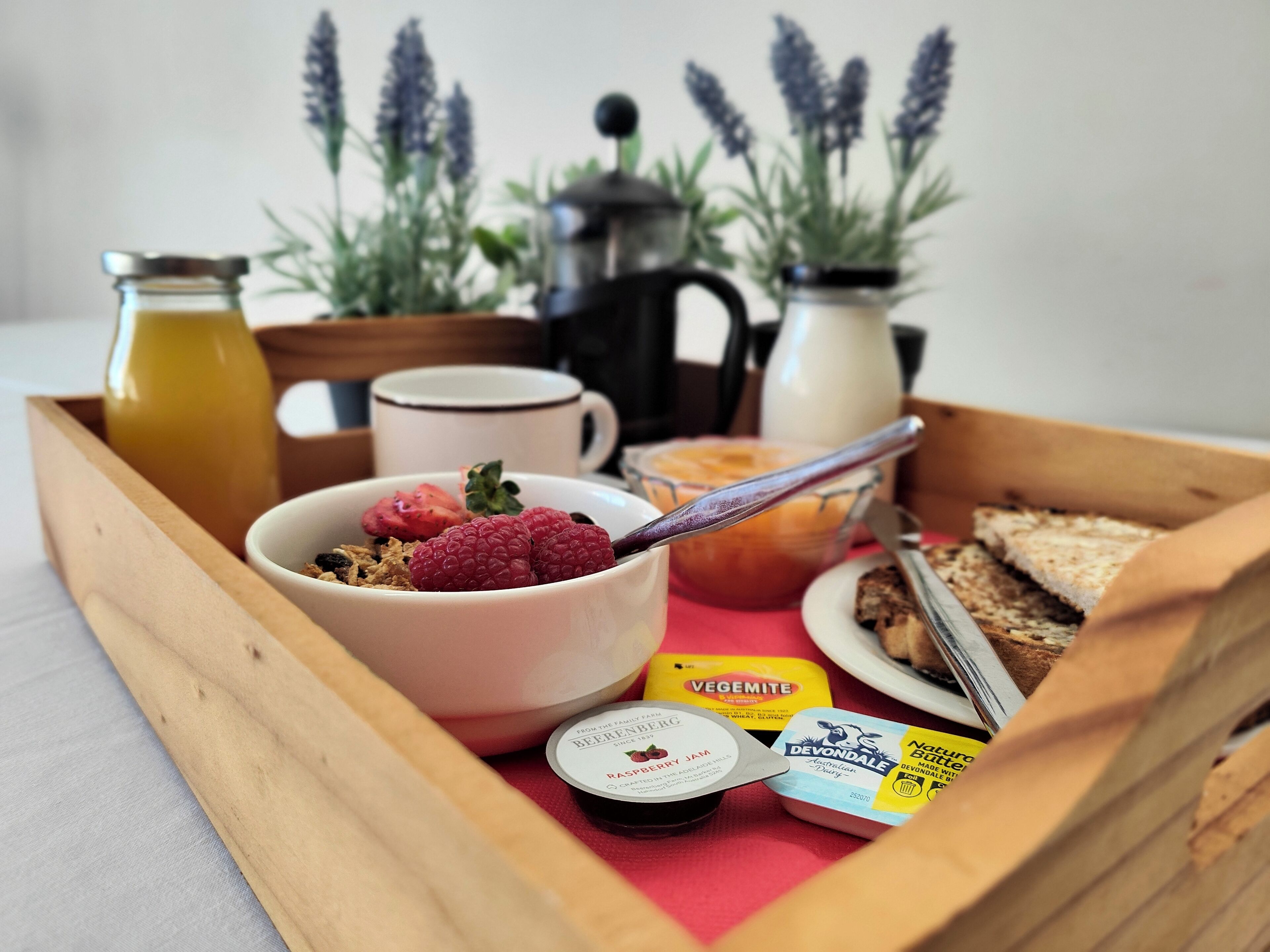 A wooden tray with breakfast items including a bowl of cereal with raspberries, a mug, a bottle of juice, a jar of jam, and a container of Vegemite.
