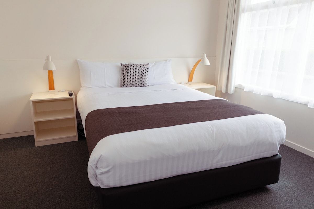 A neatly made bed with white and brown bedding, two bedside tables with lamps, and a window with sheer curtains.
