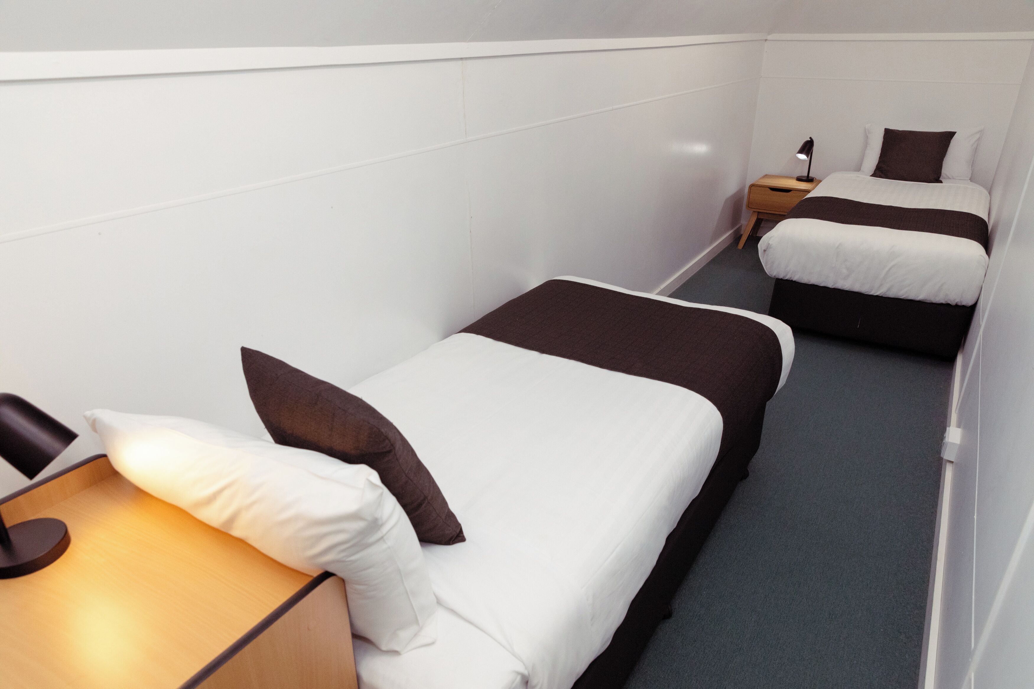A small, narrow hotel room with two single beds, each with a brown and white duvet, a bedside table with a lamp, and a small wooden nightstand.