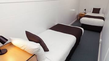 A small, narrow hotel room with two single beds, each with a brown and white duvet, a bedside table with a lamp, and a small wooden nightstand.