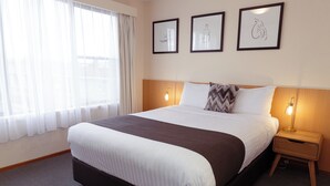 Una habitación de hotel con una cama grande, dos mesitas de noche con lámparas, un ventanal con cortinas de gasa y tres cuadros enmarcados en la pared.