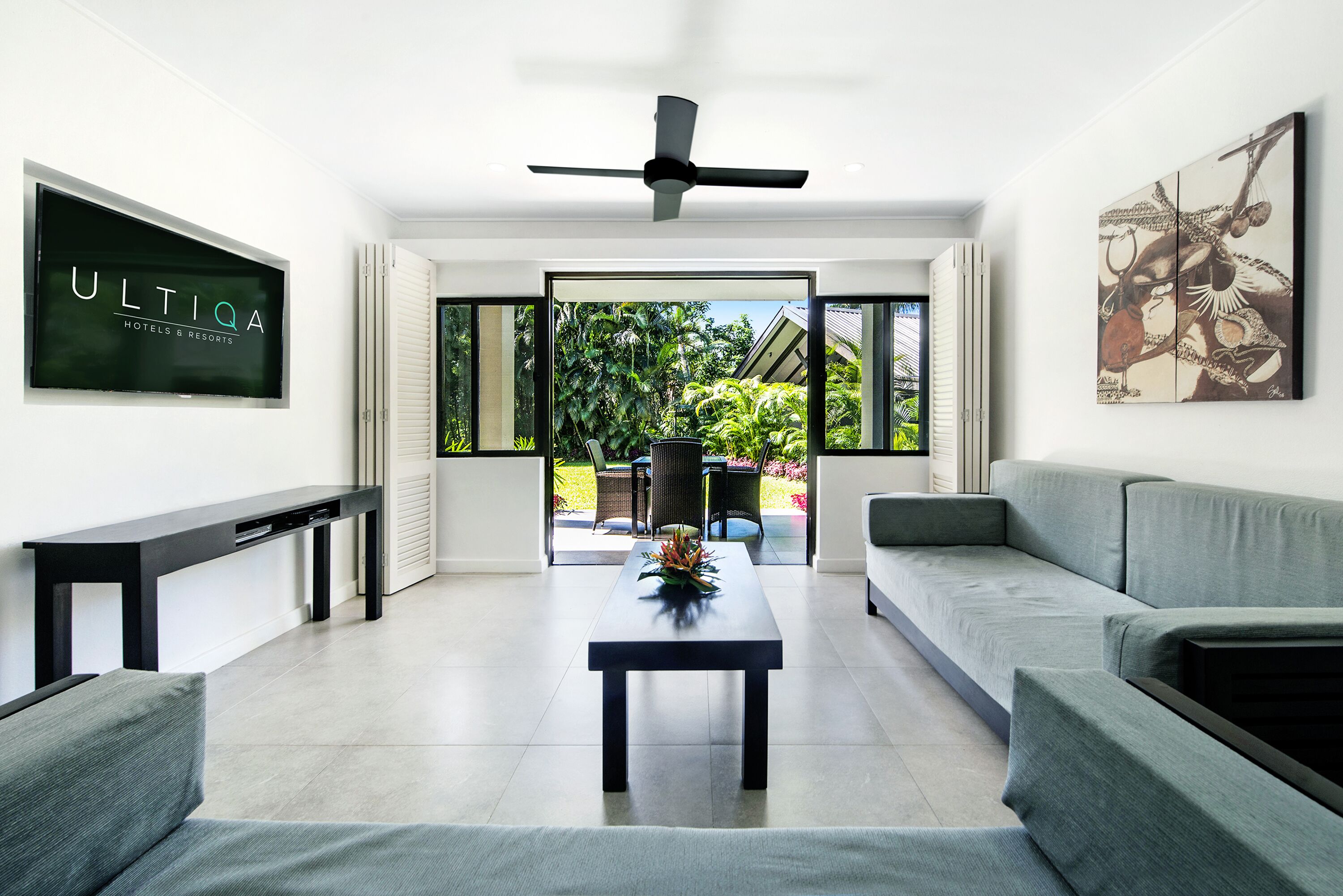 A modern living room with a flat-screen TV, a ceiling fan, a sofa, a coffee table, and a view of the outdoors.