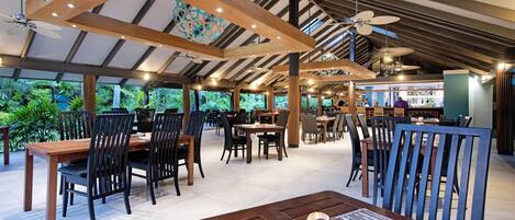 An indoor dining area with wooden tables and chairs, a bar area, and large windows offering views of greenery.