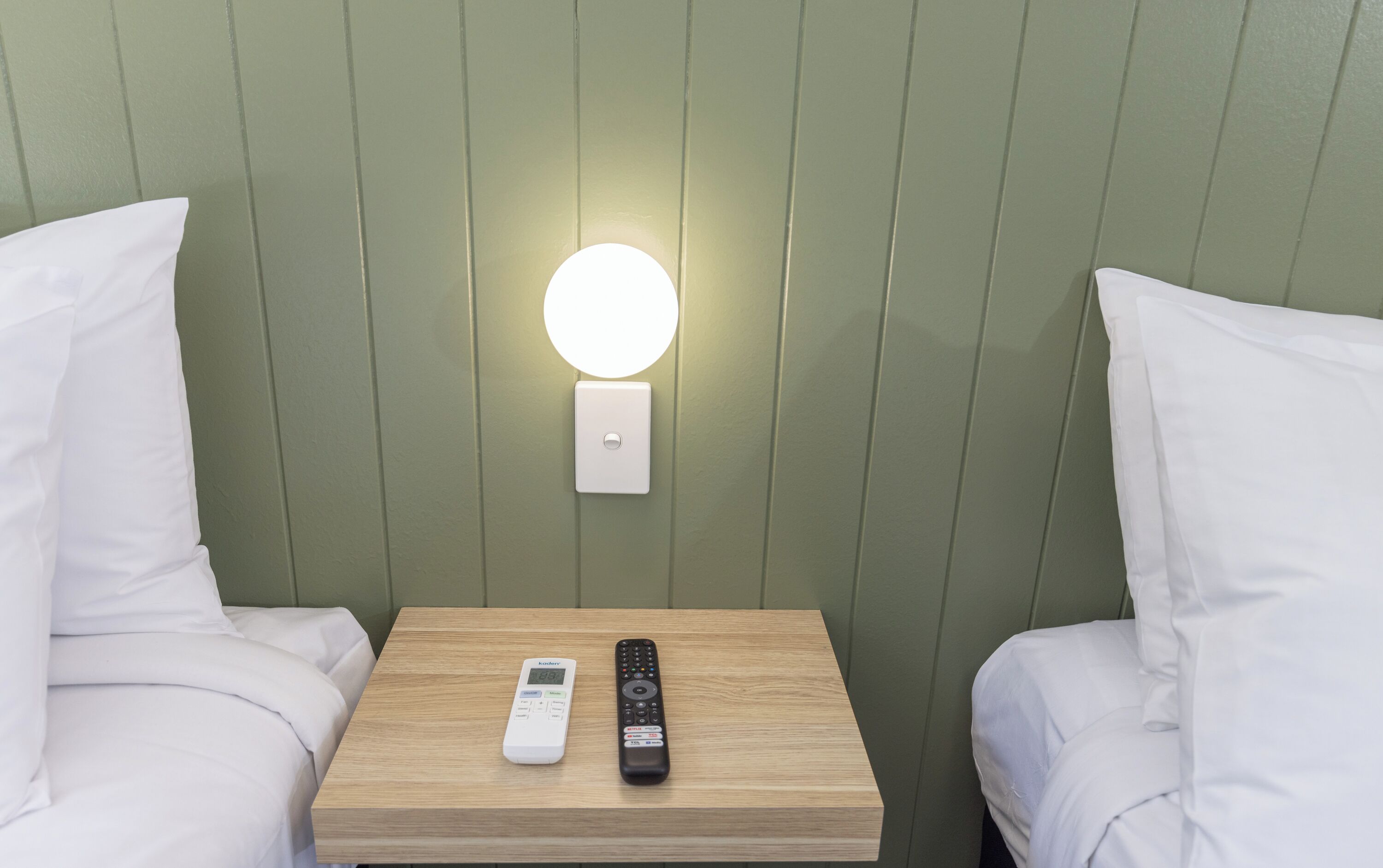 Habitación de hotel con mesita de noche, dos almohadas blancas, un control remoto y una lámpara de pared.