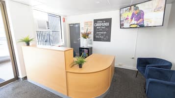 Una zona de recepción con un escritorio de madera, dos plantas en macetas, un televisor y un cartel.