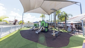 A playground with slides and a shaded area, surrounded by a fence and palm trees. - Currimundi Hotel Motel (Battery Hill)