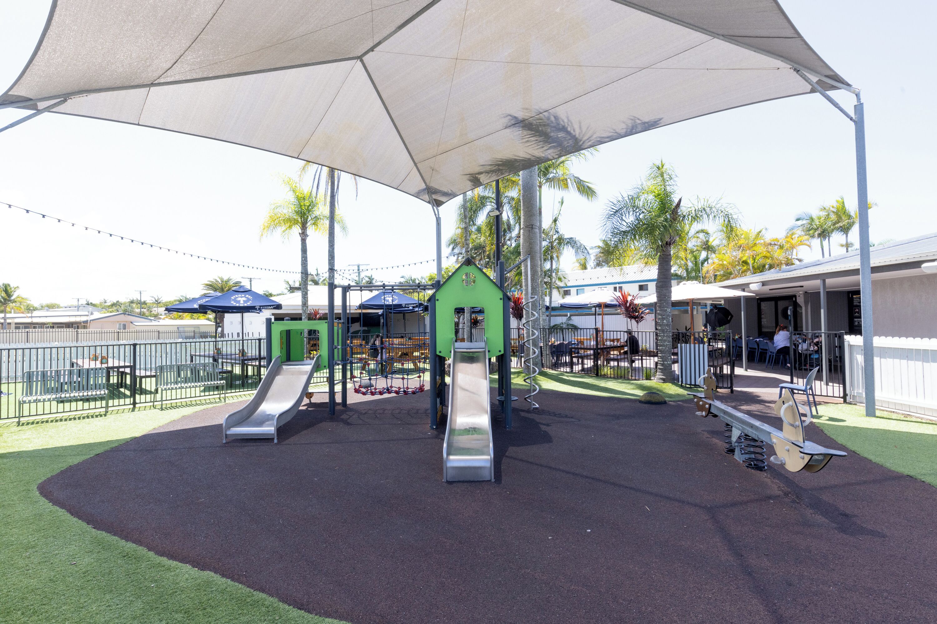 A playground with slides and swings, surrounded by a fence and palm trees.