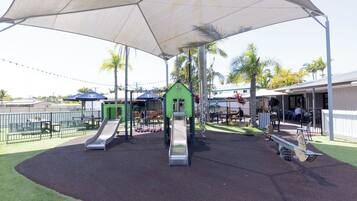 A playground with slides and swings, surrounded by a fence and palm trees.