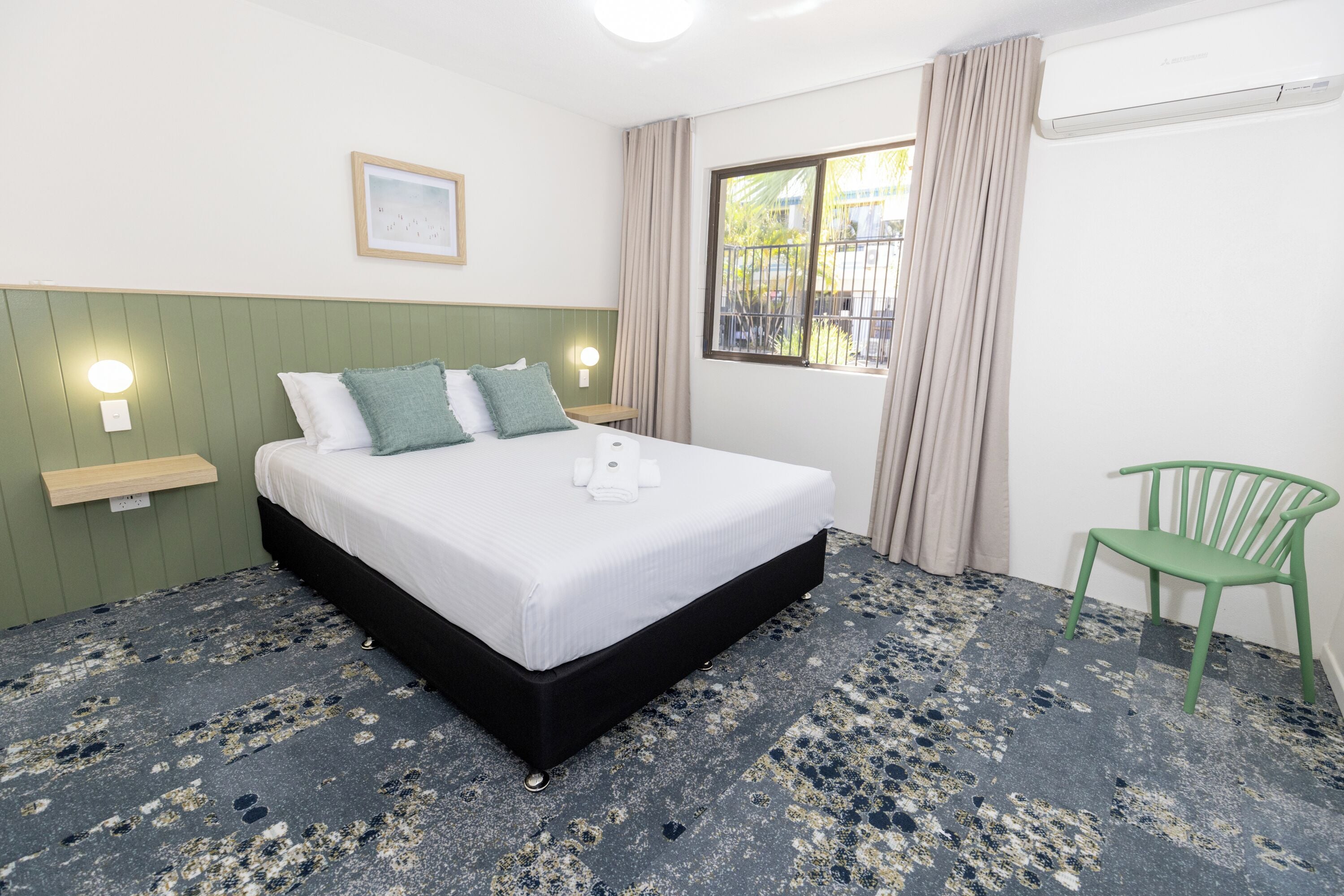 A neatly arranged hotel room with a bed, a chair, a window with curtains, a wall-mounted light, and a small table.