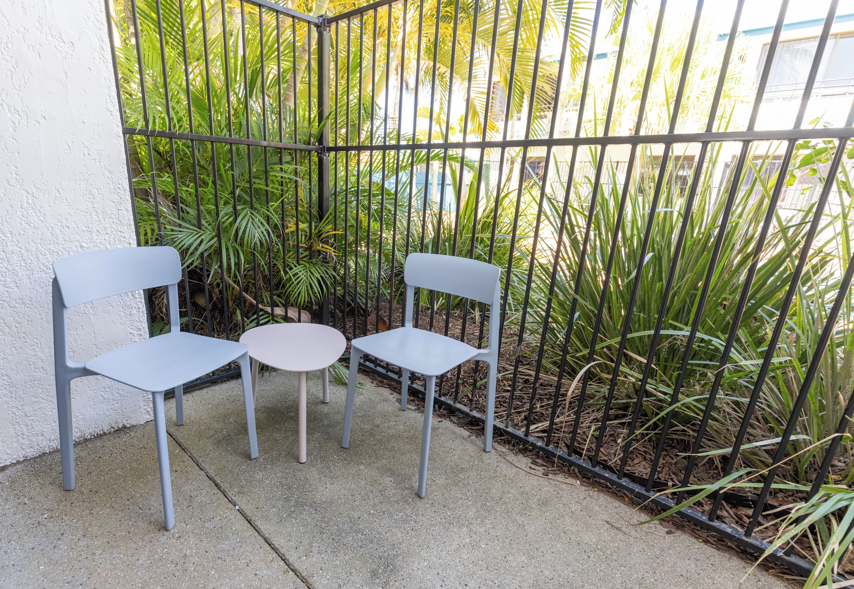 A small outdoor seating area with two chairs and a table, surrounded by a metal fence and lush greenery.