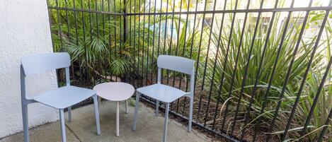 A small outdoor seating area with two chairs and a table, surrounded by a metal fence and lush greenery.