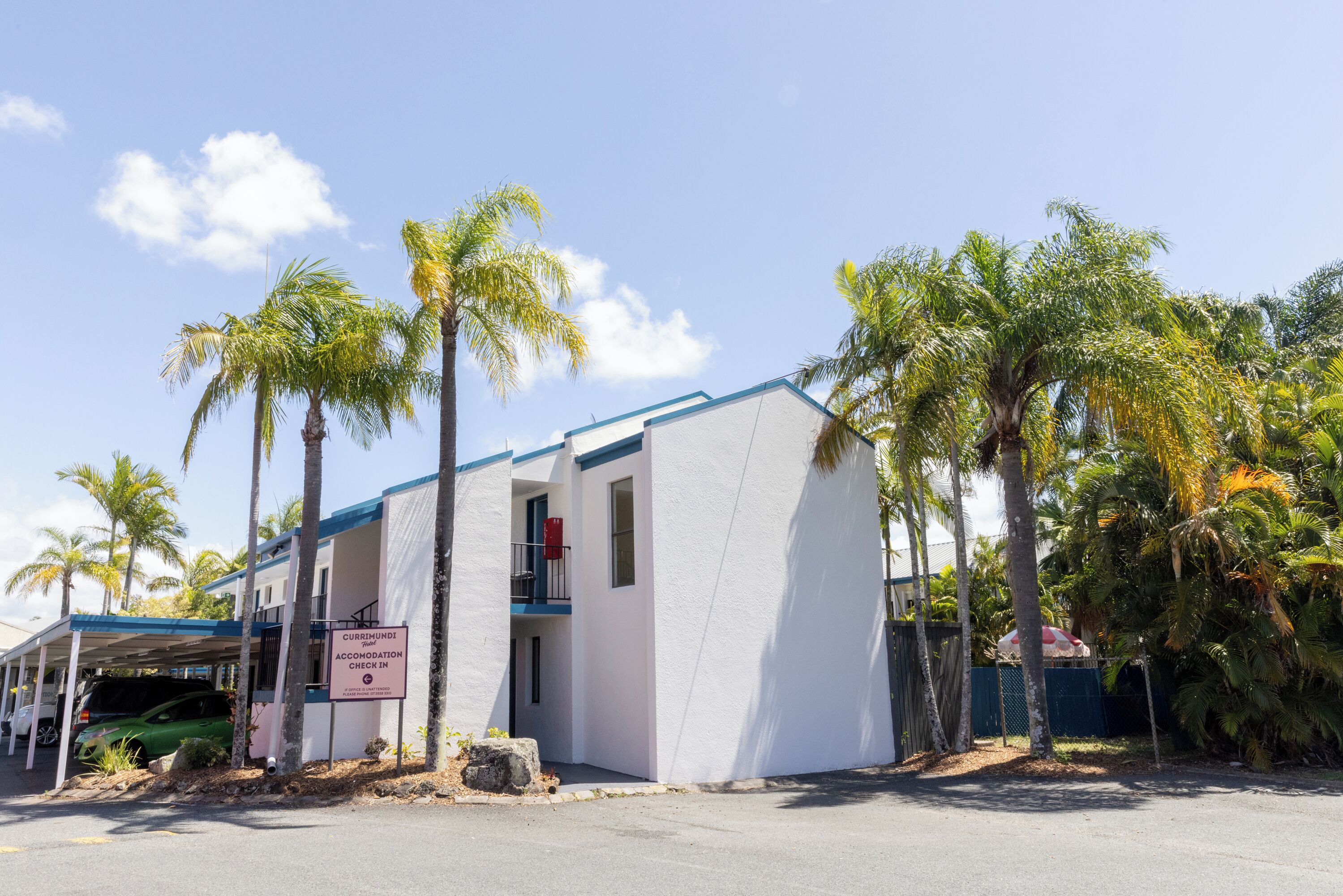 A white building with a blue roof, a sign indicating it is an accommodation check-in point, and several palm trees around it.