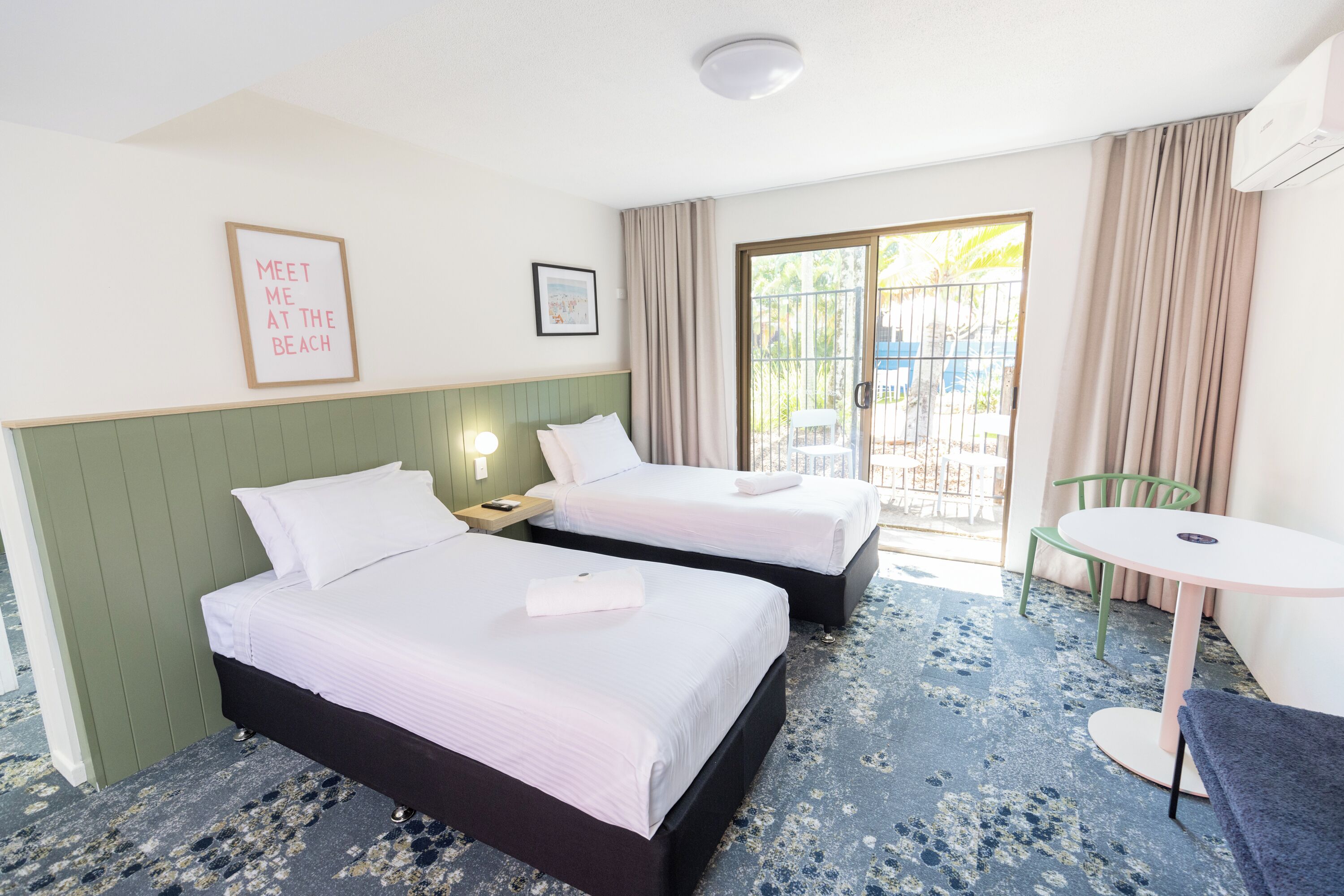 A hotel room with two beds, a small table, and a balcony with a view.