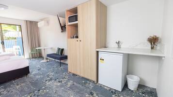 A compact hotel room with a bed, a small kitchenette, a wooden wardrobe, a desk with a chair, and a small trash bin.