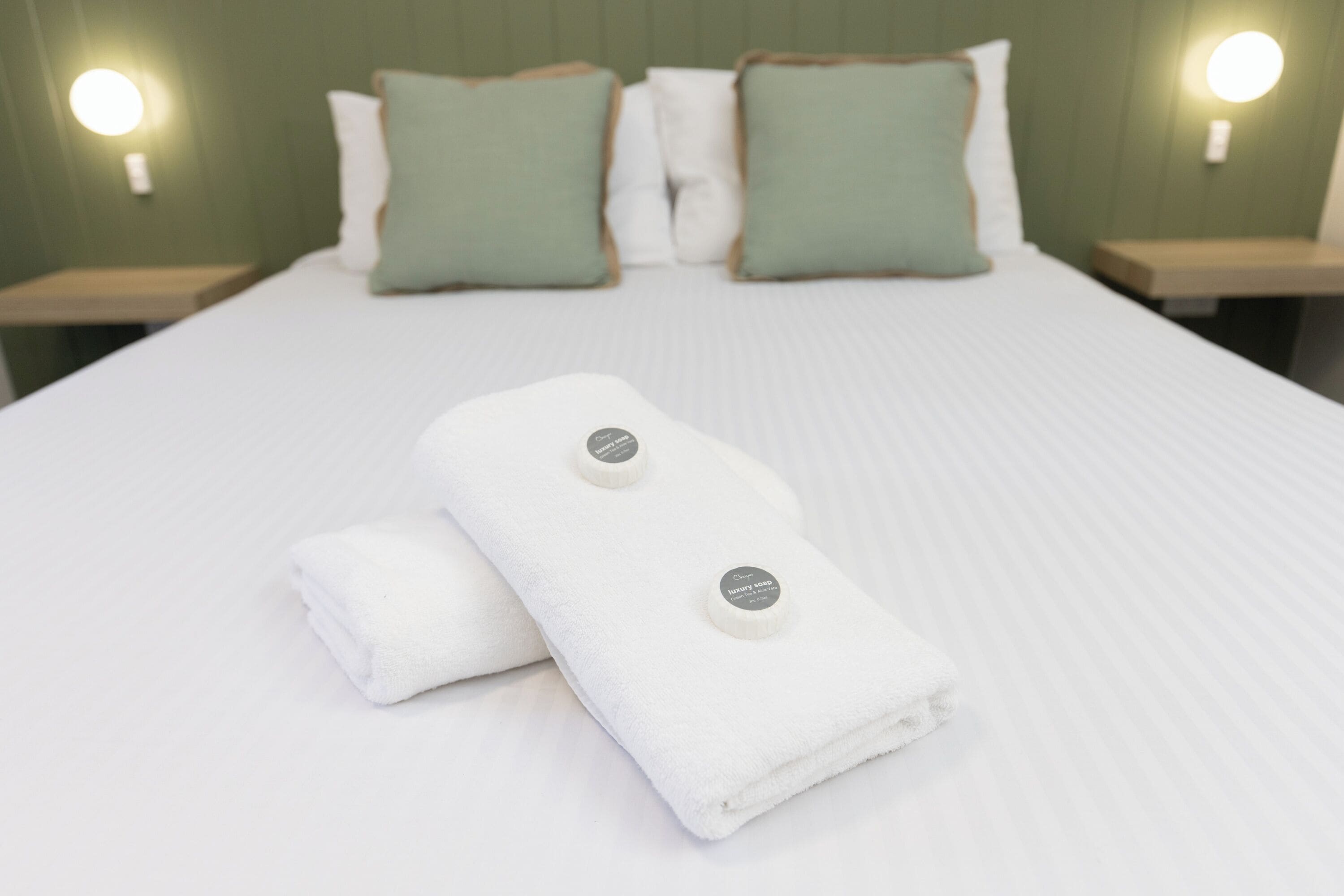 A neatly made bed with two folded towels and two pillows, one green and one white, against a green wall with two bedside lamps.
