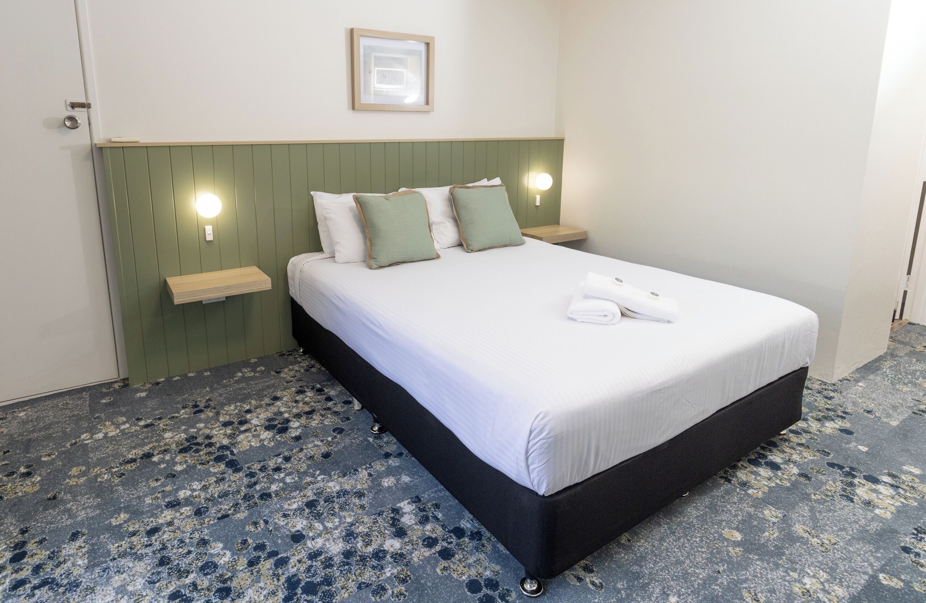 A neatly made bed with white linens and green pillows, a bedside table with a lamp, and a green headboard.