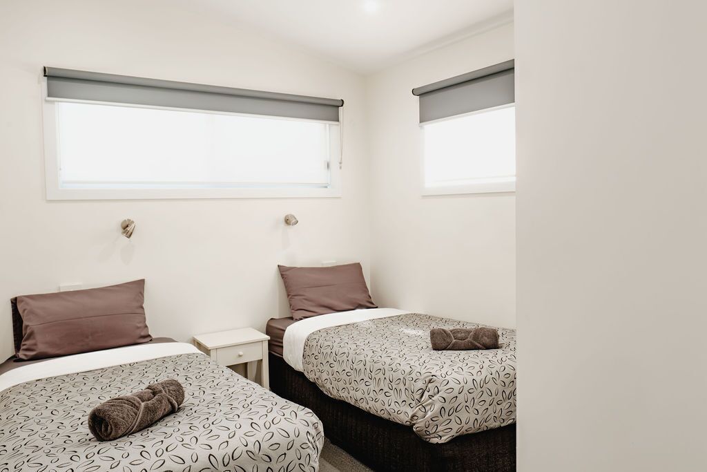 Habitación de hotel con dos camas, cada una con ropa de cama y almohadas estampadas, una mesita de noche pequeña y un ventanal con persiana enrollable.