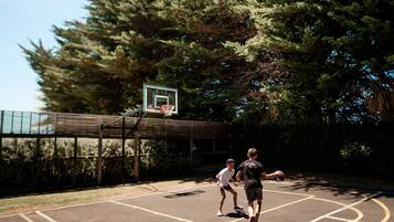 Cancha de baloncesto al aire libre con aro, rodeada de árboles y una valla.