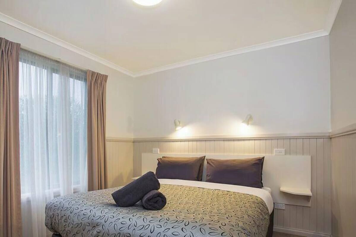 A hotel room with a bed, curtains, a headboard, and a wall-mounted light.