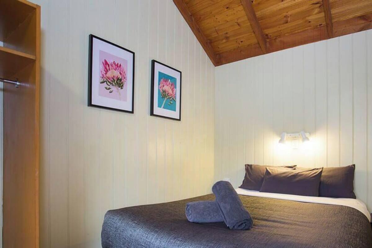 A bedroom with a bed, pillows, a nightstand, a wall-mounted light, and two framed pictures.