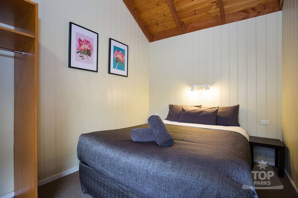 A bedroom with a bed, two framed pictures on the wall, a nightstand, and a wooden ceiling.