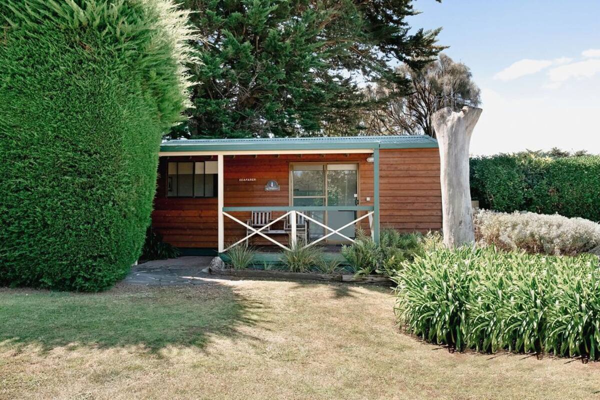 A wooden cabin with a porch, surrounded by well-maintained hedges and a large tree.