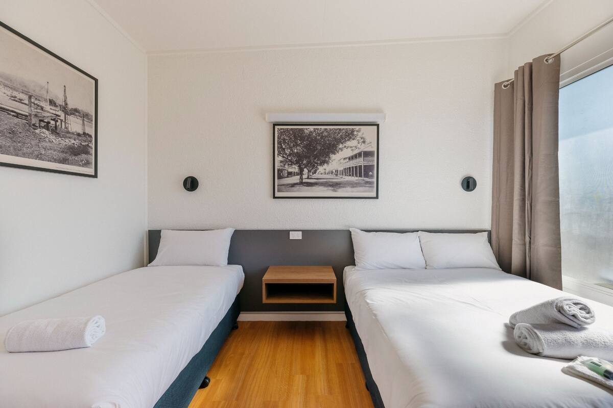 A hotel room with two single beds, a small wooden nightstand, a framed picture on the wall, and a window with curtains.