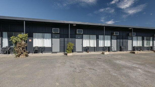 A large industrial-style building with multiple rooms, each with a window and an air conditioning unit. There are outdoor seating areas with chairs and tables, and a small tree in the foreground. - Boomerang Motor Hotel (West Mackay)