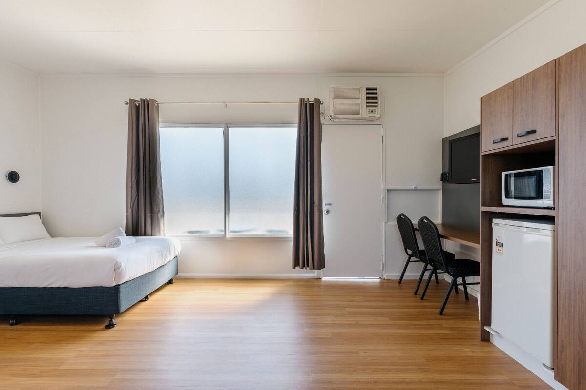 A bedroom with a bed, a desk, a chair, a microwave, and an air conditioning unit.
