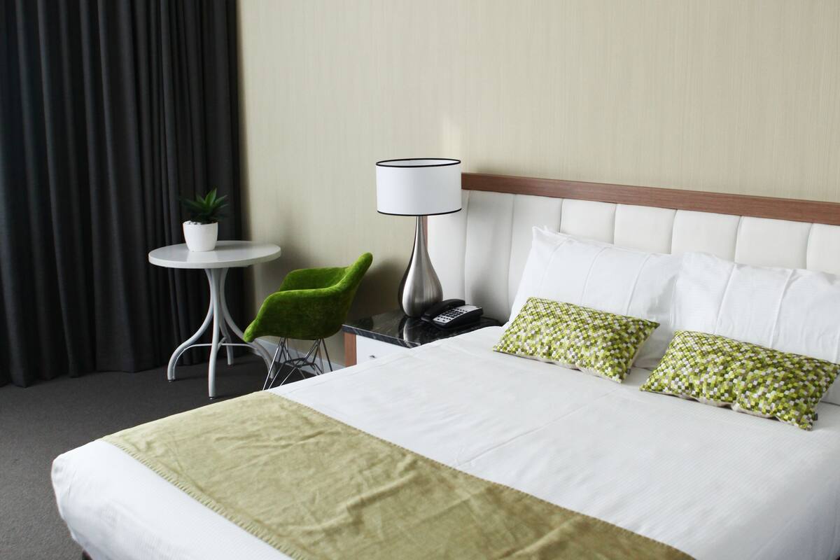 A hotel room with a large bed, a small table with a lamp, a chair, and a potted plant.