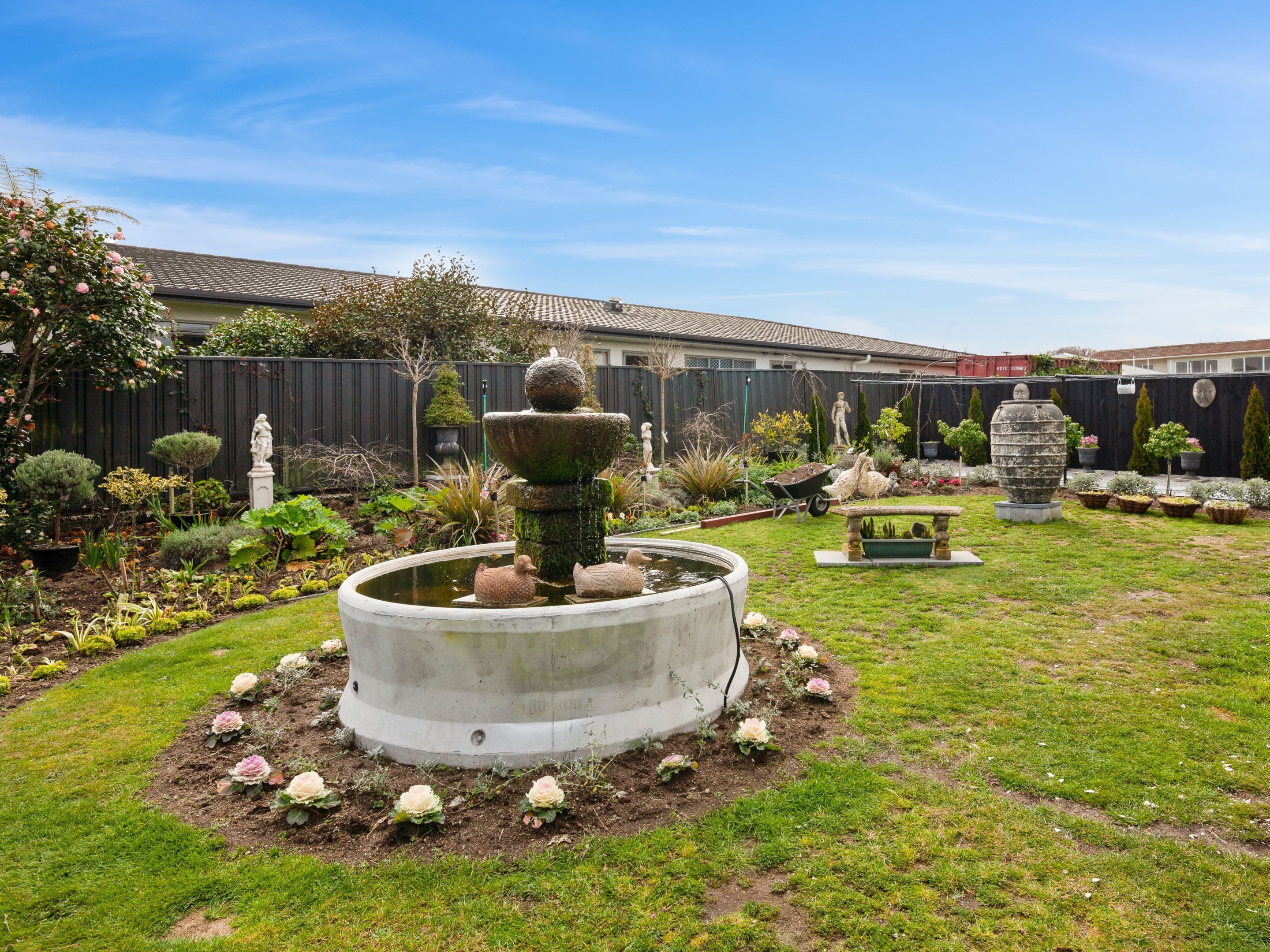 A garden with a central fountain, surrounded by various plants and a fence in the background.
