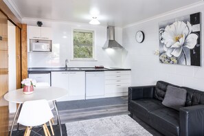 A modern kitchen with white cabinets, a microwave, and a sink. There's a dining table with chairs and a black leather sofa. A large flower painting adorns the wall. - Dunrovin Motel (Taupo)