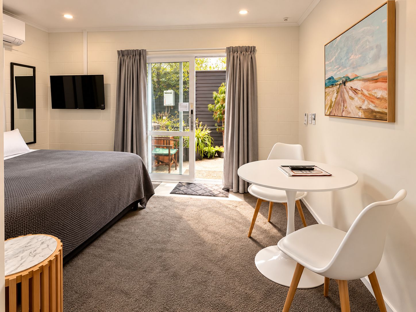 A hotel room with a bed, a round table with chairs, a painting on the wall, and a view of the outdoors through a glass door.