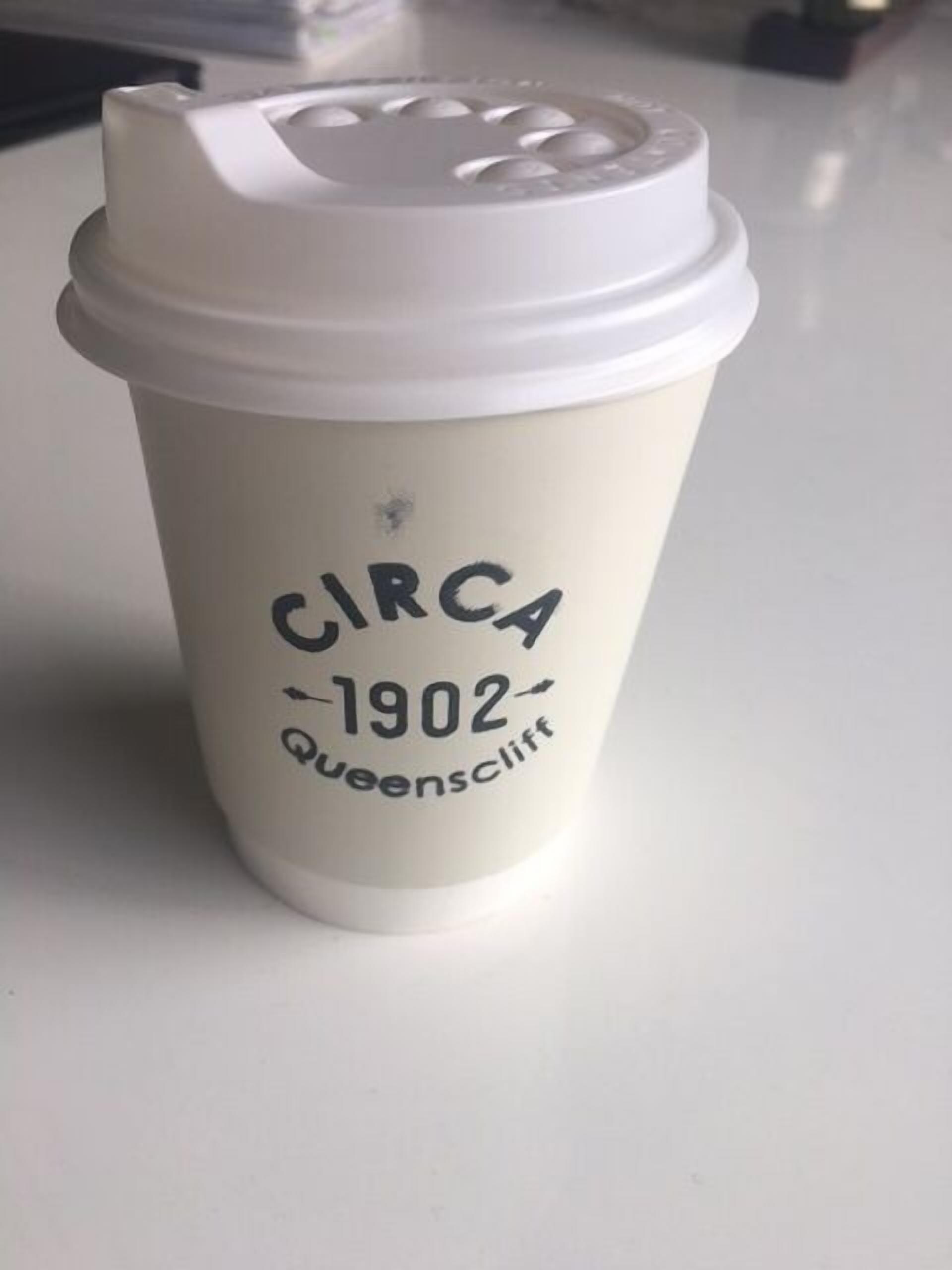 A white disposable coffee cup with the text "CIRCA 1902 Queenscliff" printed on it.