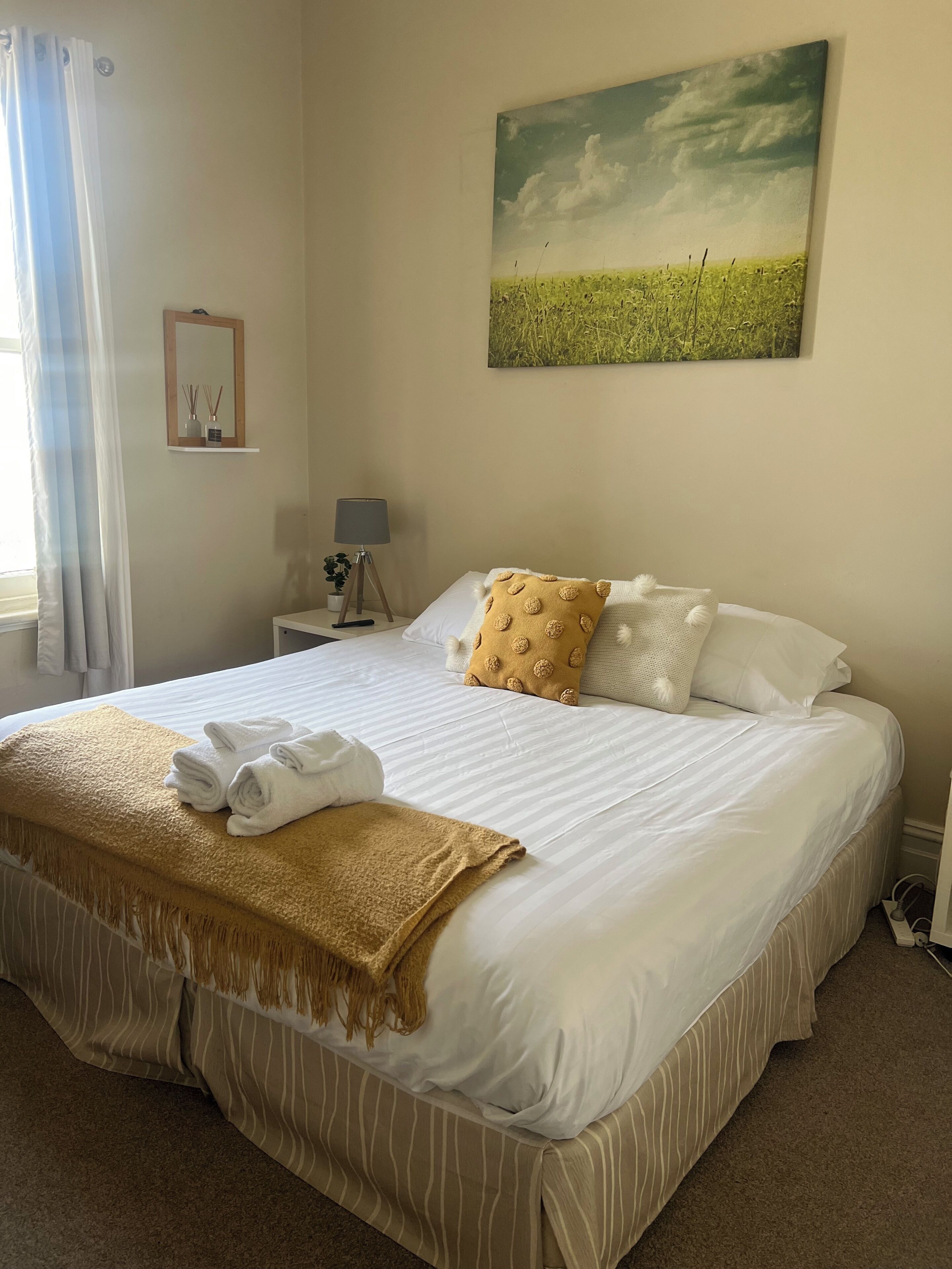 A neatly made bed with white linens and a brown throw blanket, a bedside lamp, and a framed picture of a field on the wall.