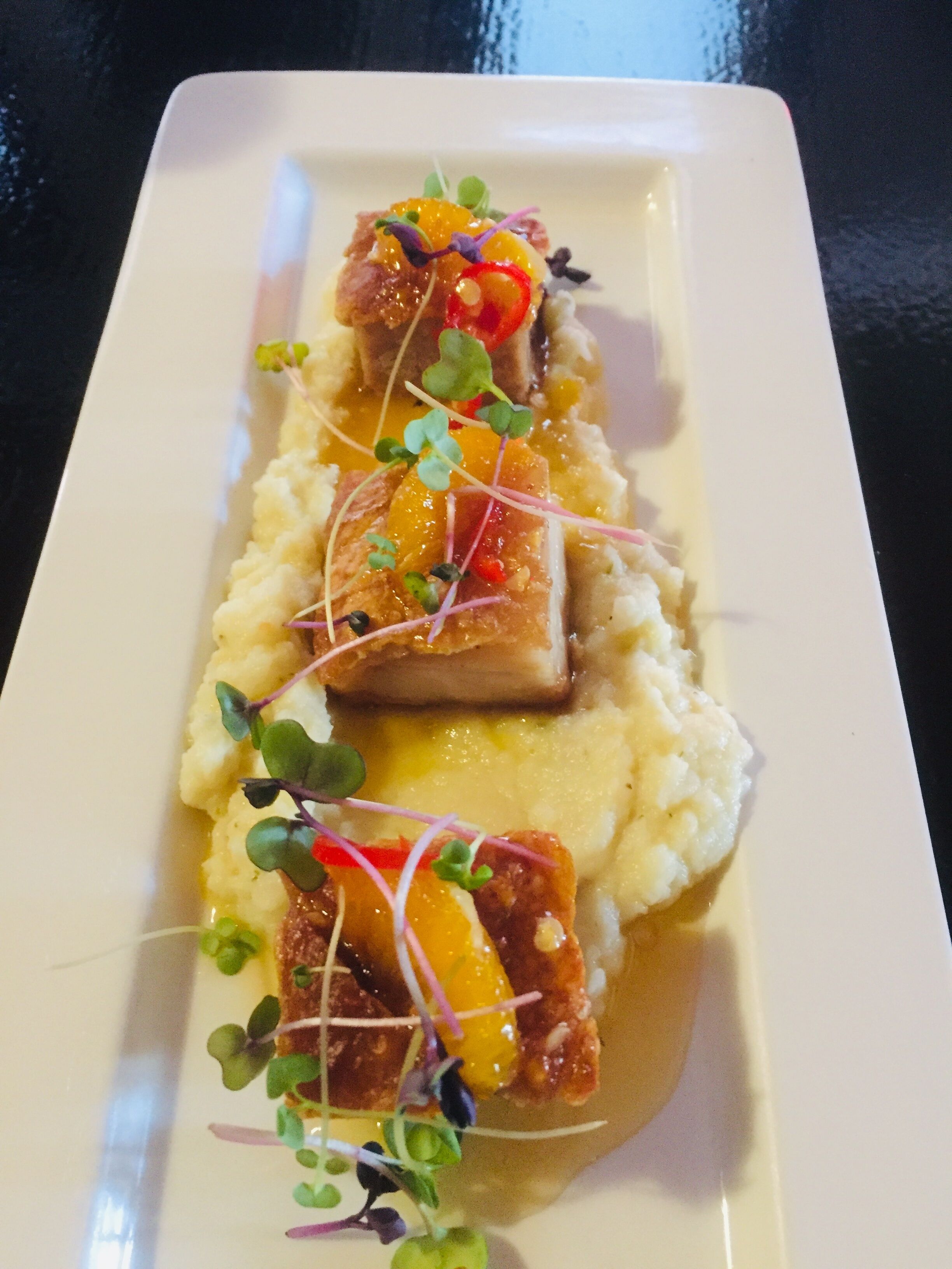 A rectangular plate with a dish consisting of mashed potatoes, a piece of meat, and garnishes.
